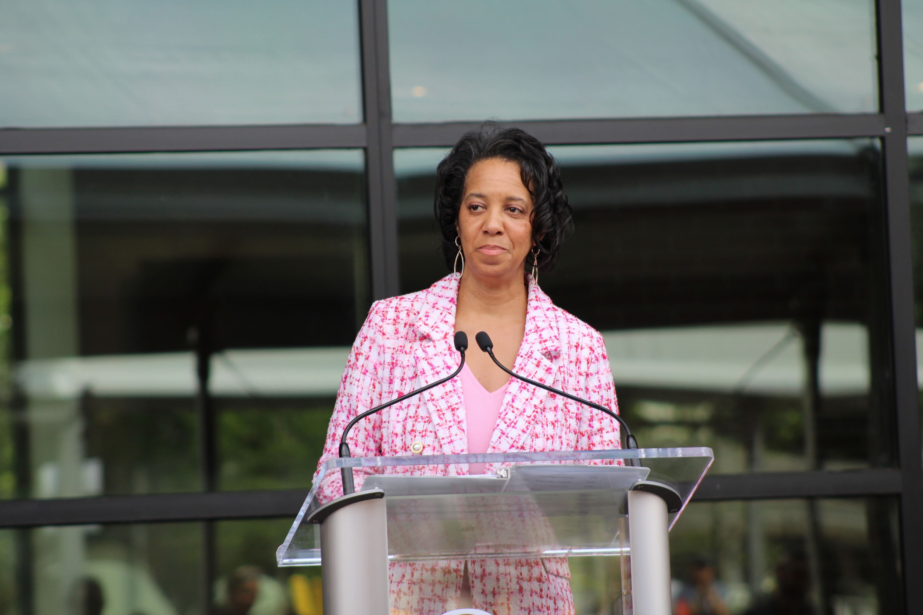 Woman in a pink patterned blazer speaking at a clear podium with dual microphones.