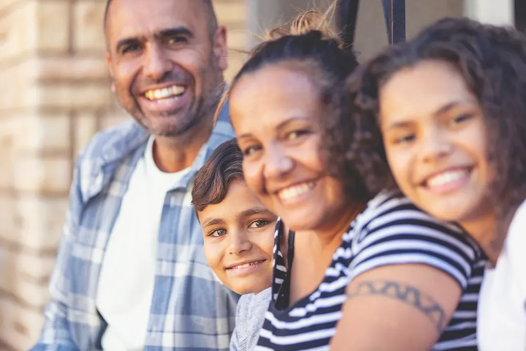 Smiling multicultural family of four sitting close together outdoors, with a blurred background.