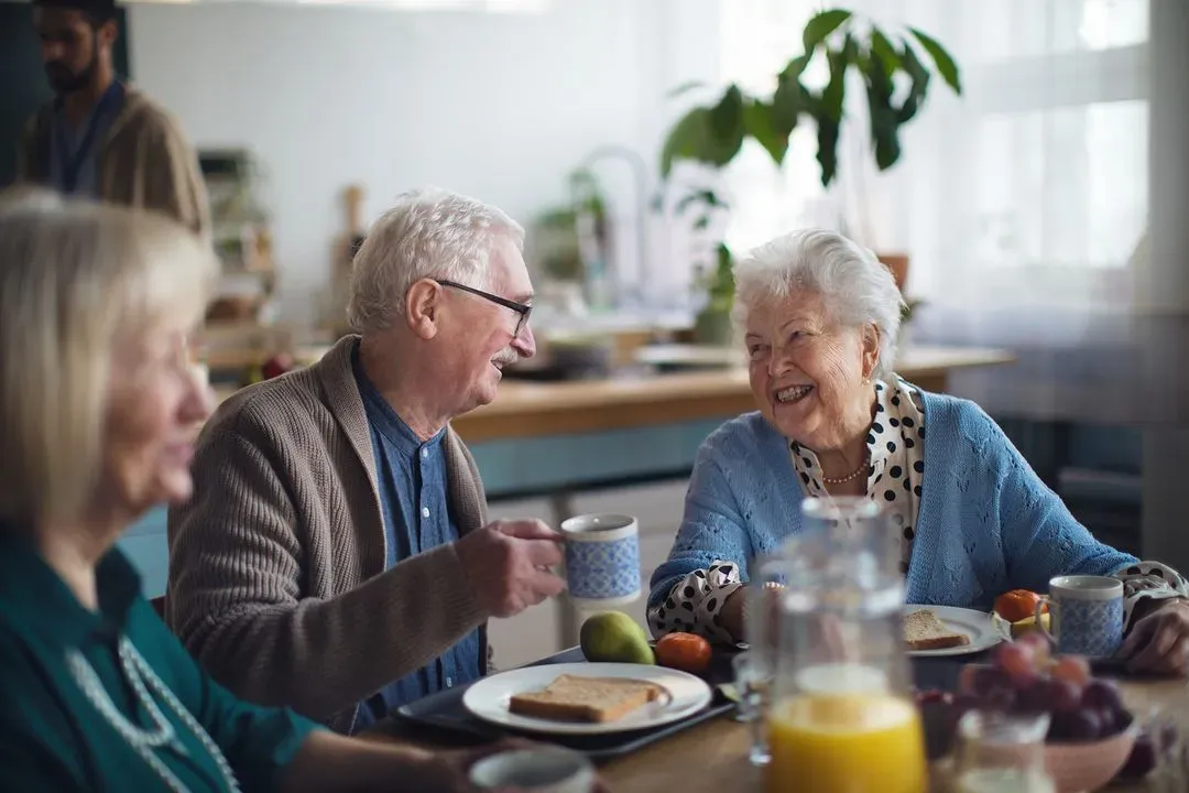 Elderly man and woman smiling and sharing a toast with coffee mugs at a breakfast table.
