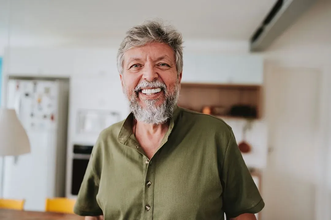 Smiling elderly man with gray hair and beard wearing a green shirt in a modern kitchen.