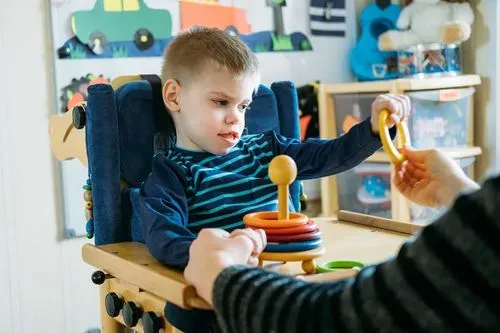 Young boy in a wheelchair playing with a ring stacking toy assisted by an adult.