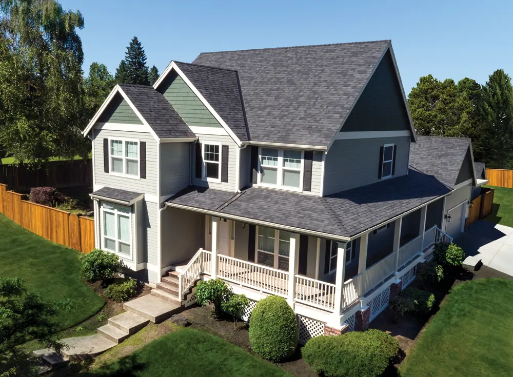 Two-story gray house with dark gray shingle roof, white trim, front porch, and landscaped yard with green grass and bushes.