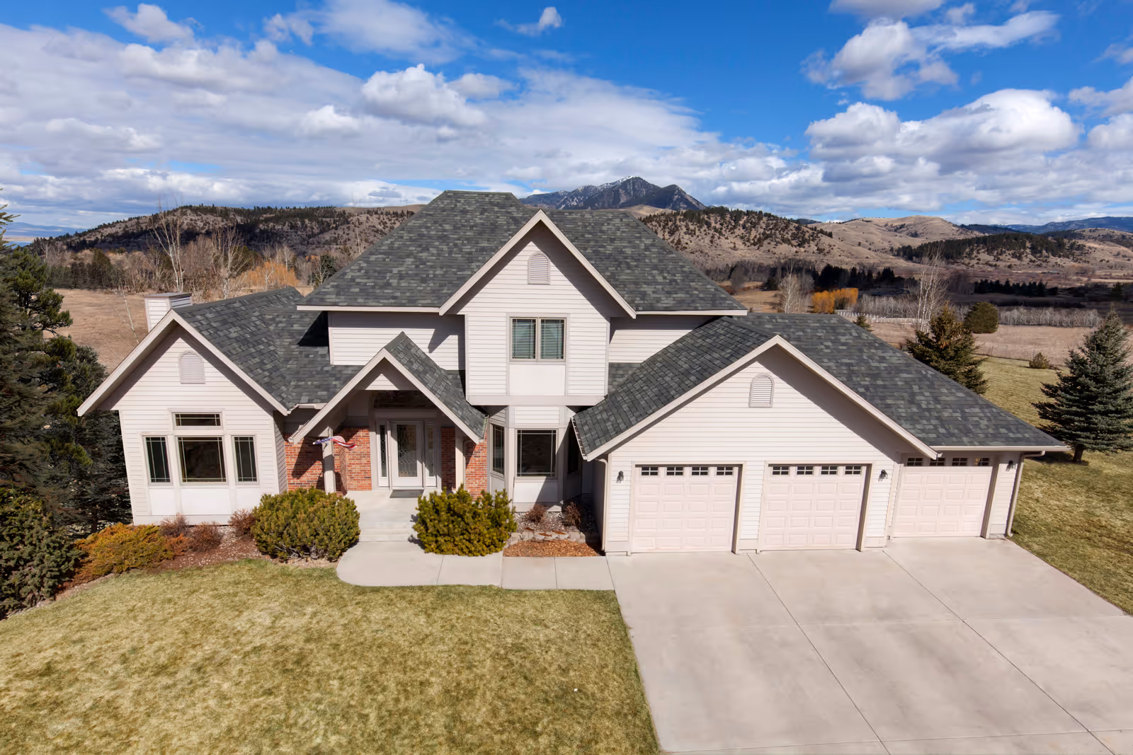 Two-story suburban house with gray shingled roof, three-car garage, and mountain landscape in the background.