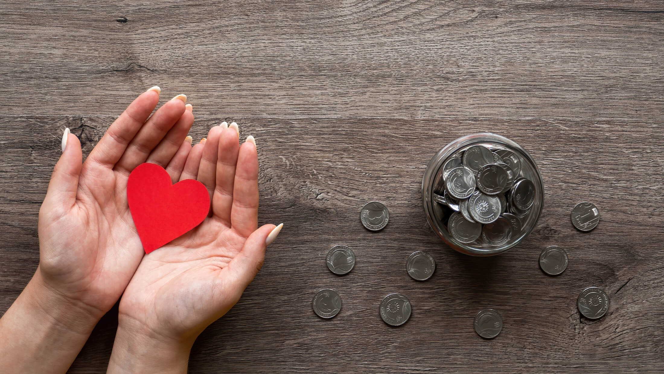 A person holding a red heart in their hand. A jar of coins sits next to them on the table