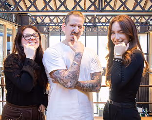 Trois adultes souriants posant avec la main sous le menton dans un espace intérieur lumineux avec des poutres apparentes.