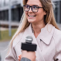 Jeune femme souriante portant des lunettes, interviewée avec un microphone en extérieur.