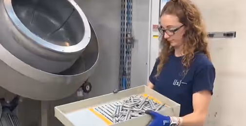 Une femme porte un plateau contenant plusieurs pièces métalliques devant une machine industrielle.