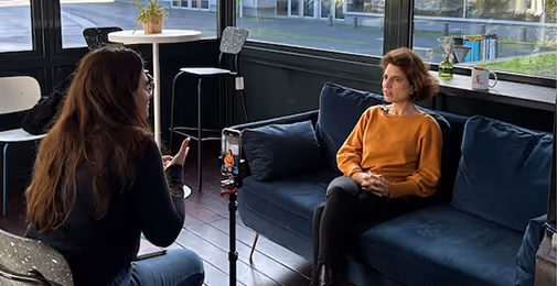 Une femme assise dans un canapé bleu lors d'une interview avec une autre femme tenant une caméra sur trépied.