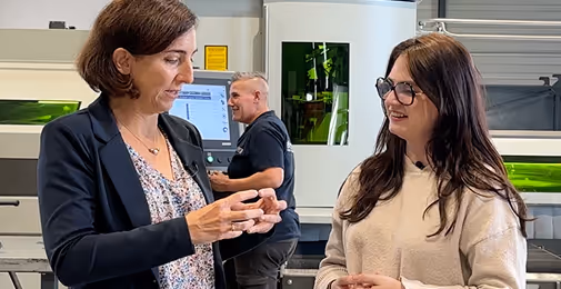 Deux femmes discutant dans un environnement industriel avec un homme en arrière-plan devant une machine.