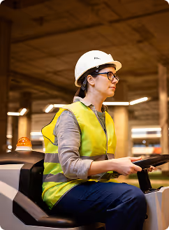 Femme portant un casque de sécurité et un gilet réfléchissant jaune, assise dans un véhicule de chantier.