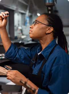 Une femme portant des lunettes et un tablier bleu examine attentivement une petite pièce dans un atelier.