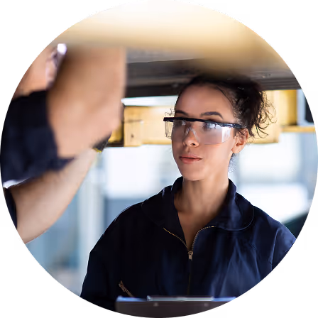 Femme portant des lunettes de sécurité et une combinaison de travail, inspectant un mécanicien sous un véhicule.