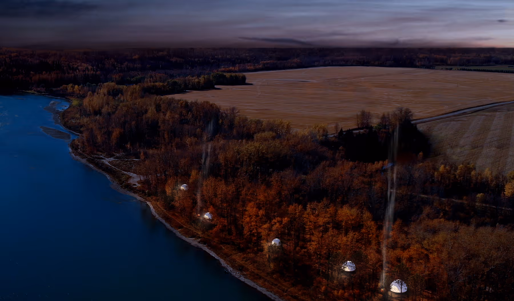 Aerial view of a lakeside forest with autumn foliage and five illuminated white dome tents emitting smoke.