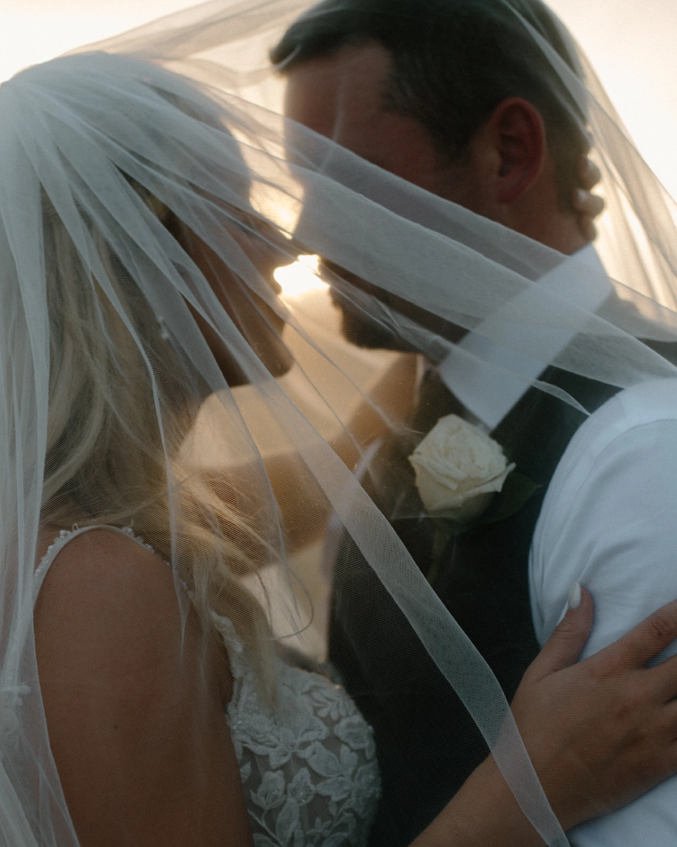 A bride and groom stand closely under a veil with soft, warm light filtering through. The groom wears a white rose boutonniere. Romantic and intimate mood.