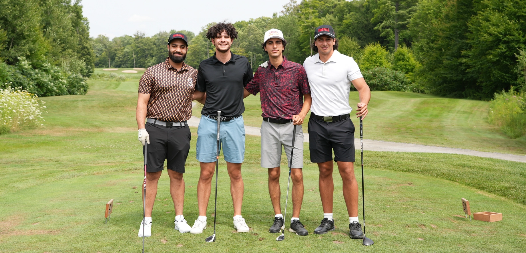 Four men stand on a lush golf course, each holding a club at a Fireball Wiskey golf event. They smile warmly, dressed in casual golf attire, with trees lining the fairway behind.
