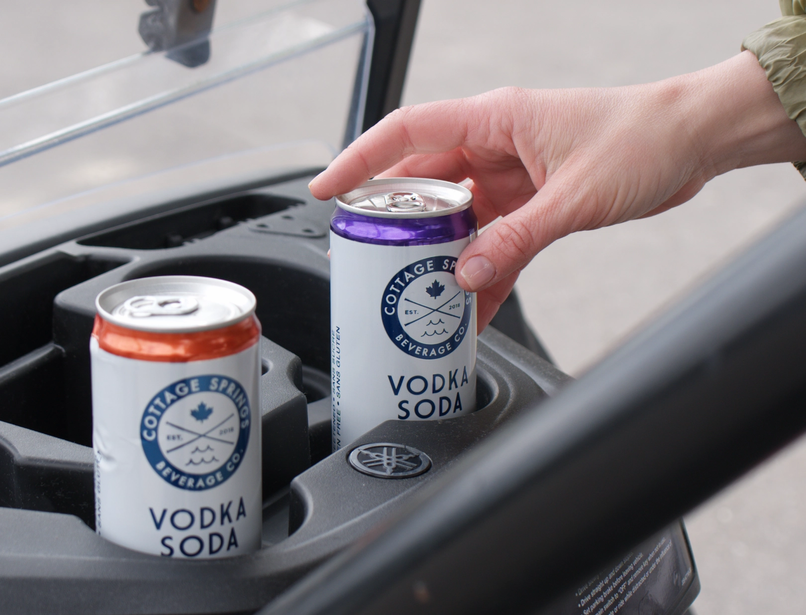 A hand reaches for a can of Cottage Springs Vodka Soda inside a golf cart cup holder, with another can of the same drink beside it.