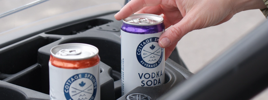 A hand reaches for a can of Cottage Springs Vodka Soda inside a golf cart cup holder, with another can of the same drink beside it.