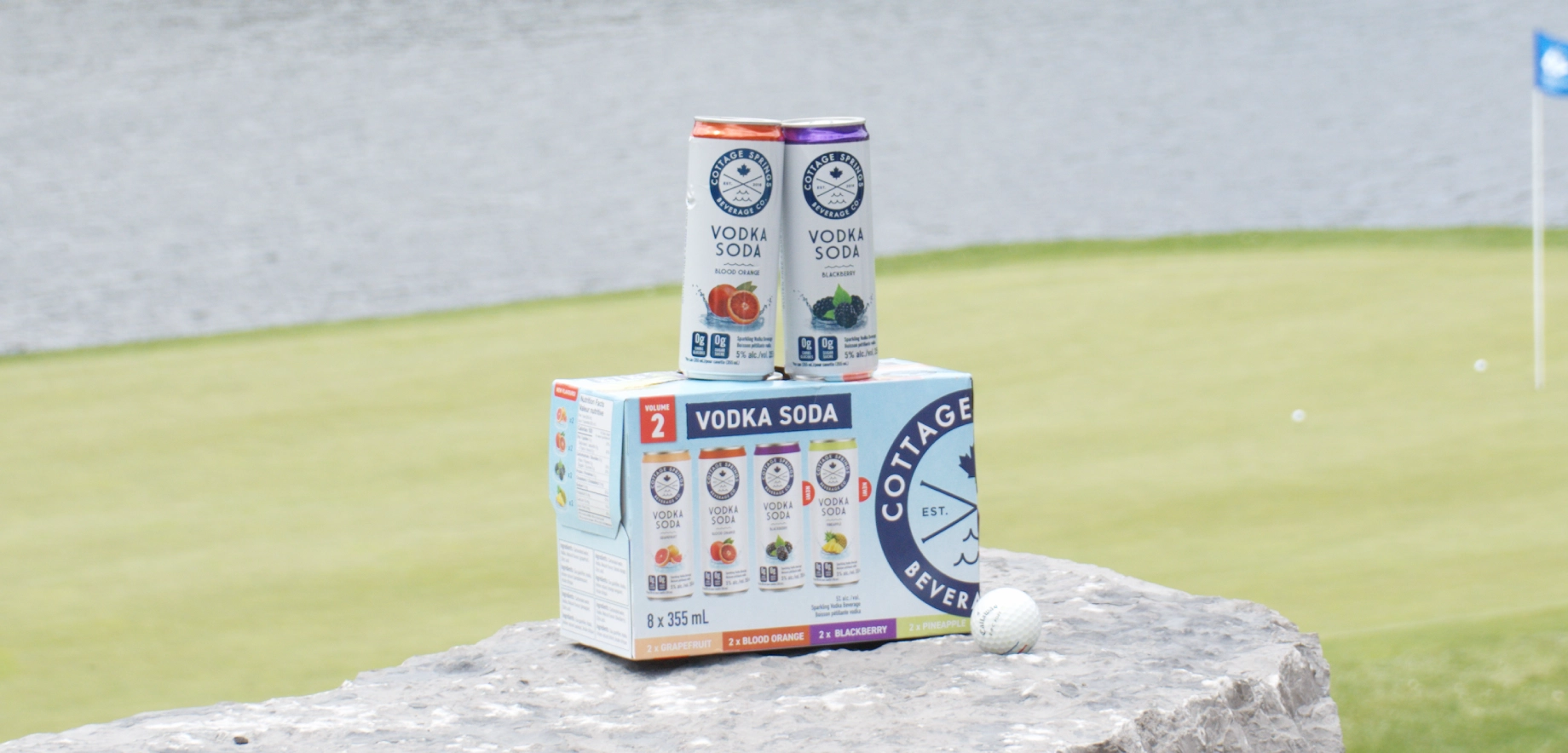 Two cans of Cottage Prings Vodka Soda sit atop a pack on a stone ledge, overlooking a golf course. A golf ball rests nearby, with a calm water view in the distance.