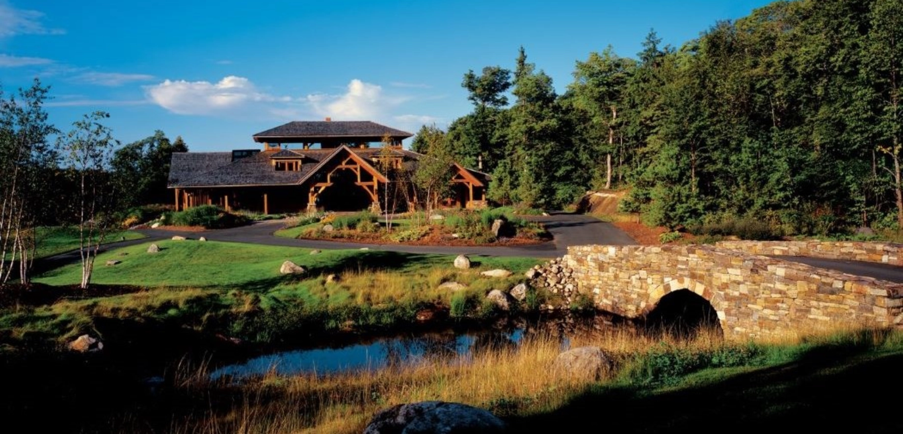 The rustic Ridge at Manitou lodge with wooden beams is surrounded by lush trees under a blue sky. A stone bridge crosses a tranquil pond in the foreground.
