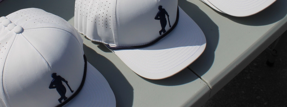 White baseball caps with perforated panels and the Gentlemens Golf logo on the front in black are neatly arranged on a light-colored table, creating a sporty vibe.