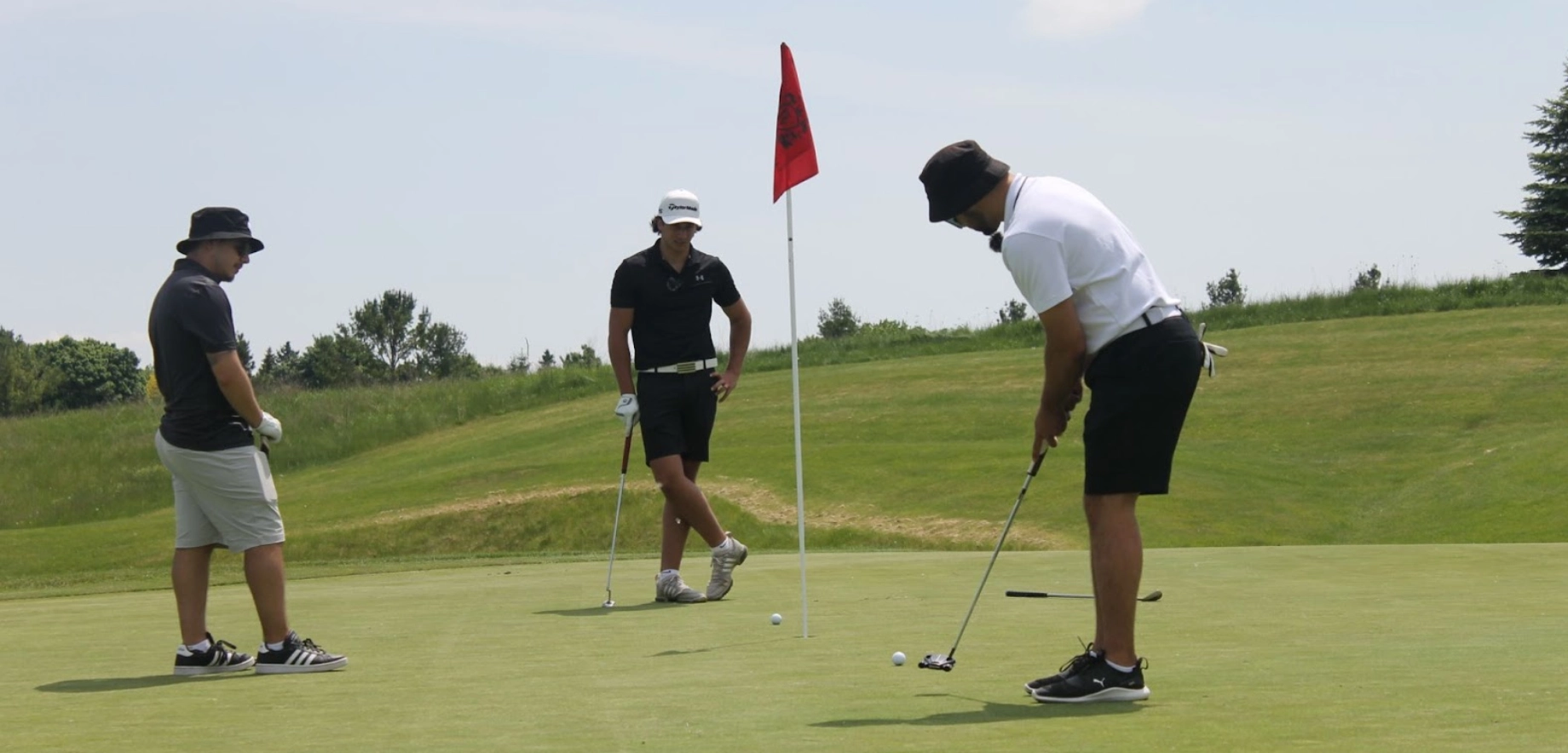 Three golfers on a green, with one lining up a putt near a red flag. The scene is sunny, with a relaxed and focused atmosphere.