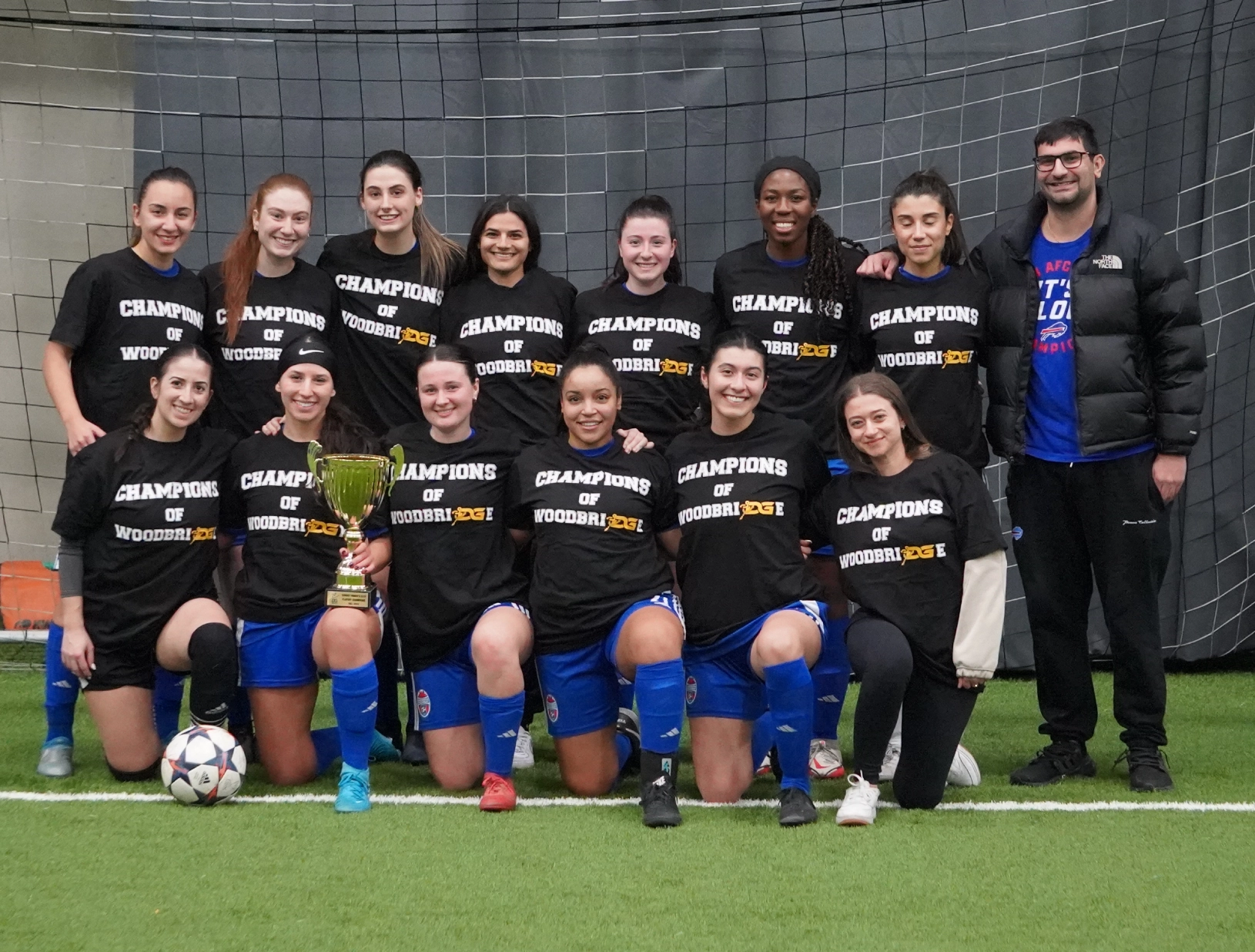 A women's soccer team poses triumphantly in front of a net at the DG Sports indoor soccer field, wearing "Champions of Woodbridge" shirts and holding a trophy. There's a joyous, celebratory mood.