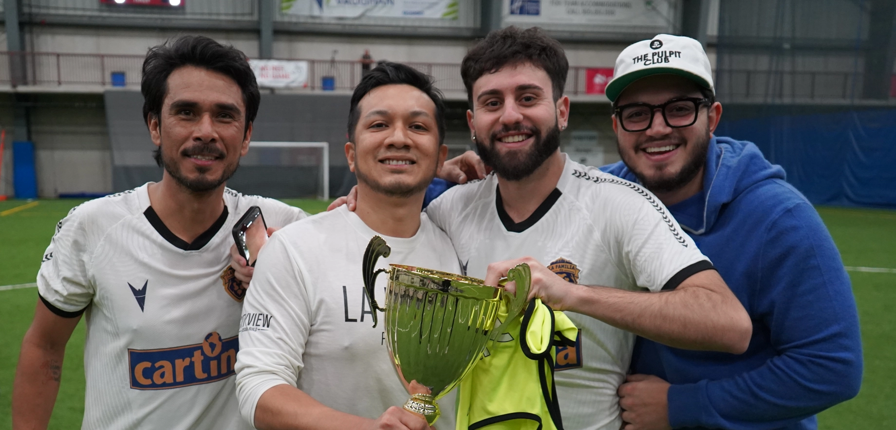 Four men in soccer jerseys and casual wear smile and hold a trophy on an indoor field. The atmosphere is joyful and celebratory.