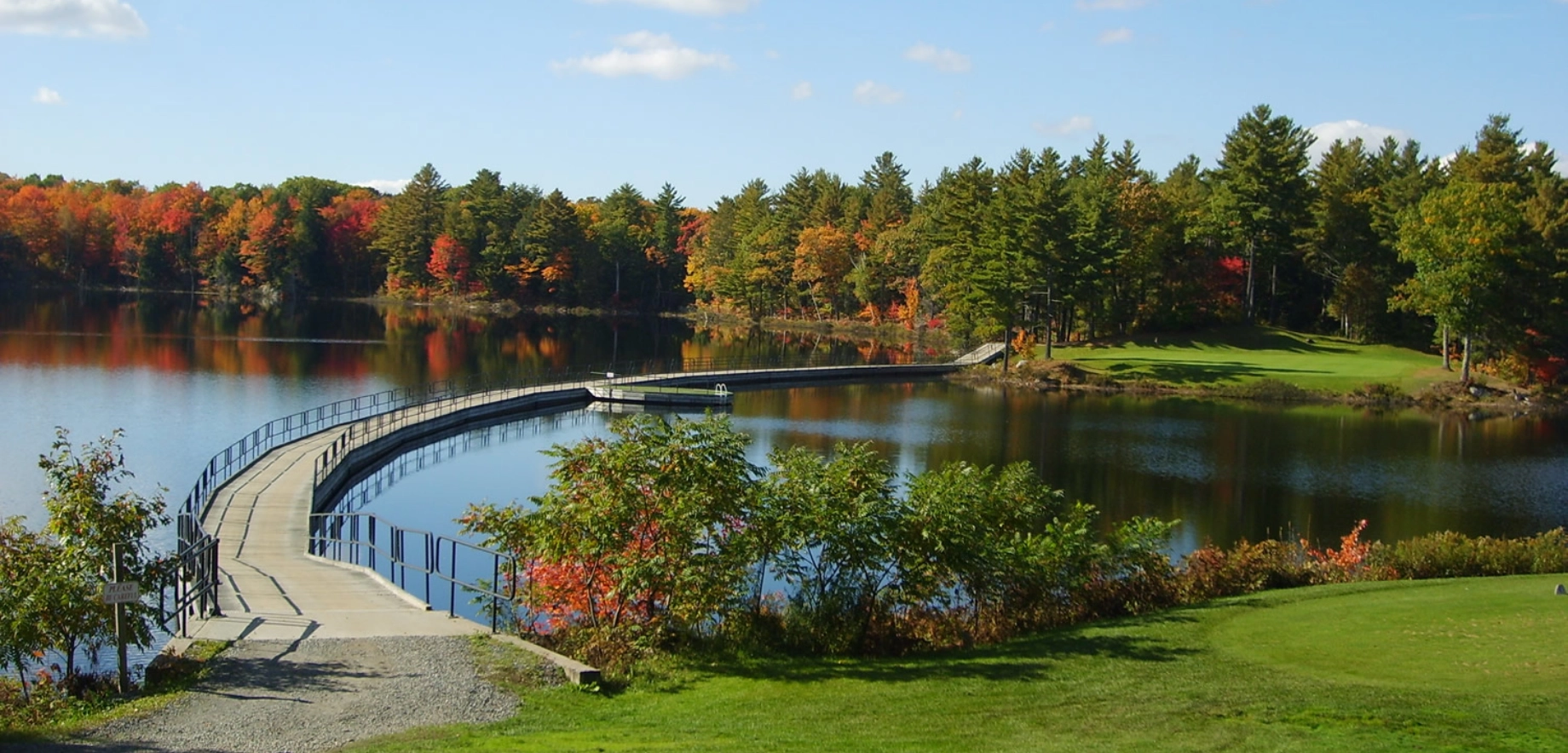 Curved bridge over calm lake at the Seguin Valley golf coursse reflects autumn trees under a clear sky. Lush green foreground contrasts with vibrant fall foliage, creating a serene scene.