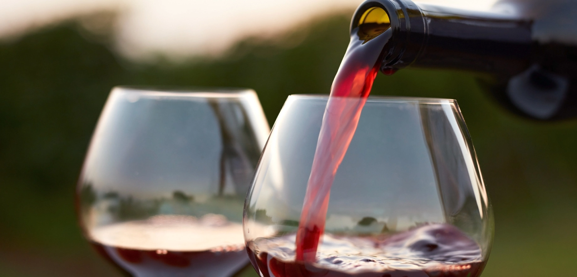 Red wine is being poured from a bottle into one of two glasses outdoors. The setting sun casts a warm glow, creating a relaxed and elegant ambiance.