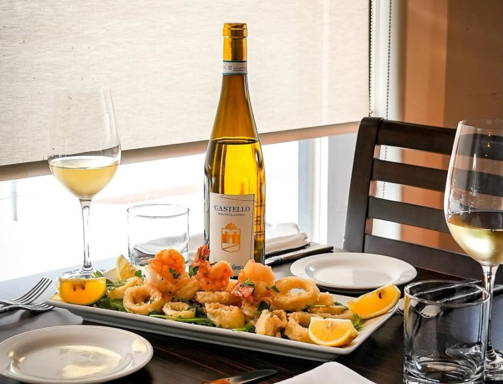 A bottle of white wine with two glasses on a table, alongside a seafood platter with fried calamari and shrimp. The setting conveys elegance and warmth.