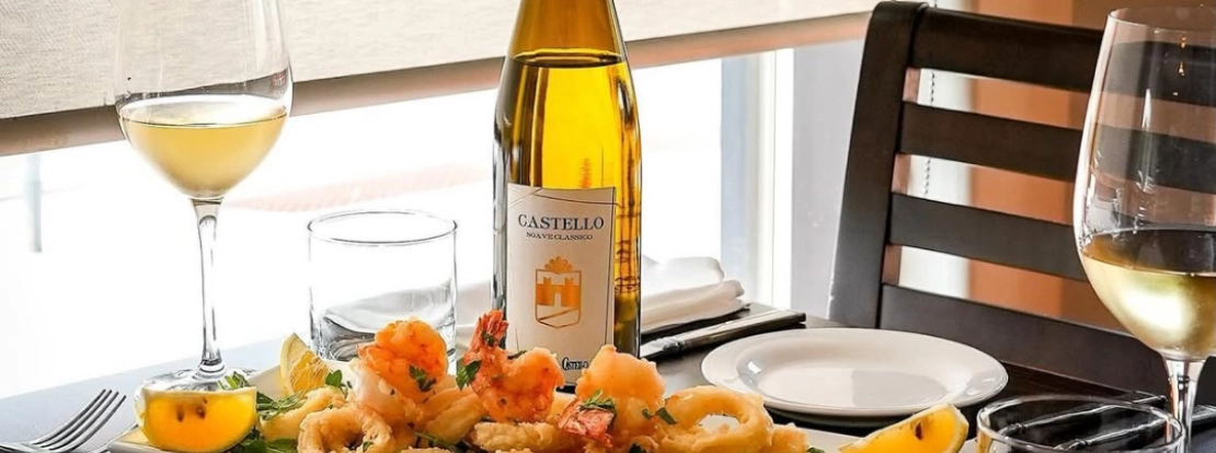 A bottle of white wine with two glasses on a table, alongside a seafood platter with fried calamari and shrimp. The setting conveys elegance and warmth.