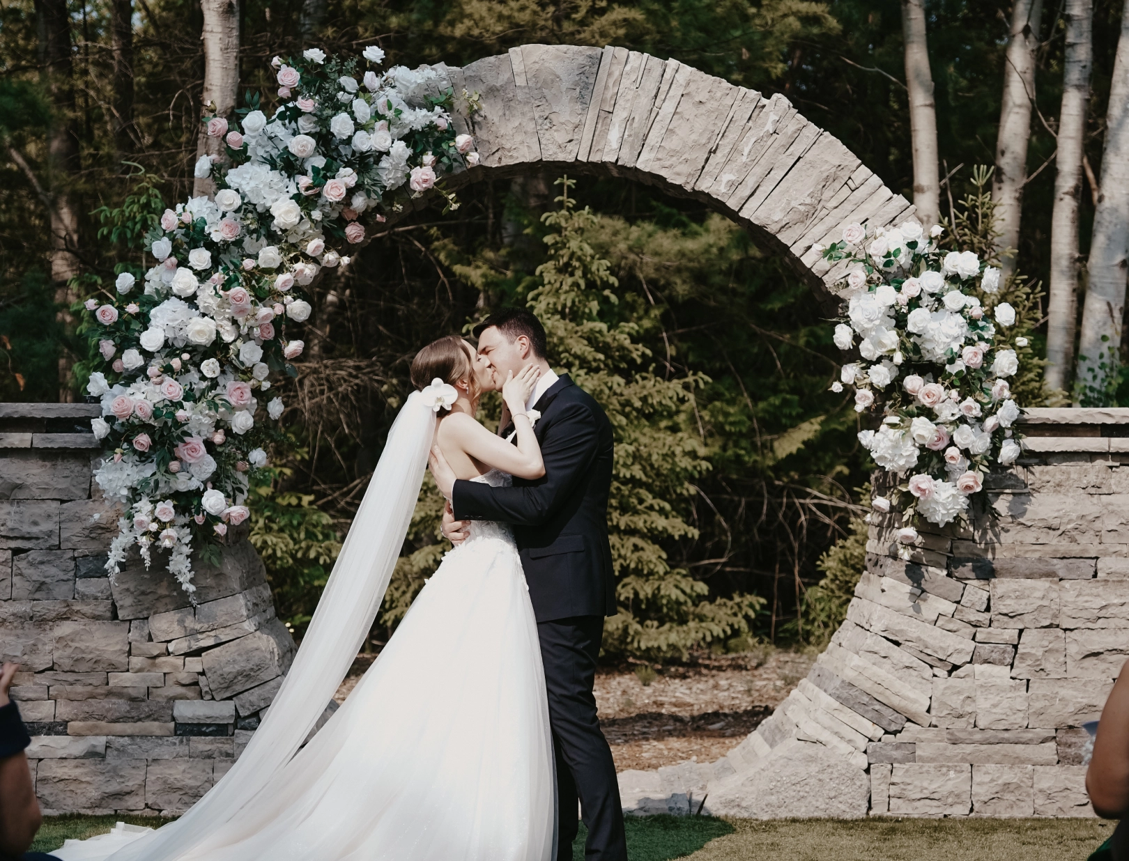 A bride and groom kiss under a circular stone arch adorned with white and pink flowers. The scene is romantic, set against a forest backdrop.