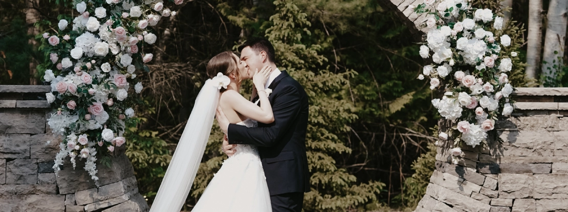 A bride and groom kiss under a circular stone arch adorned with white and pink flowers. The scene is romantic, set against a forest backdrop.