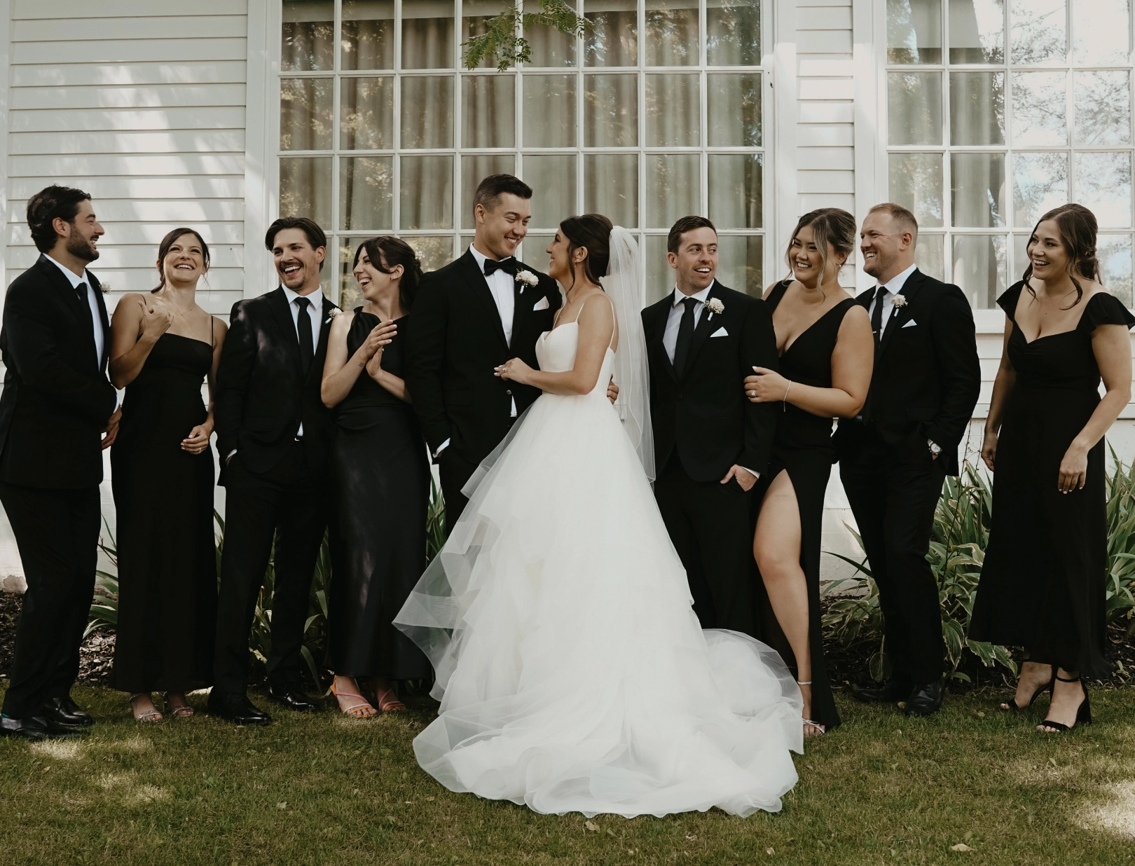 A joyful wedding party stands in front of a white building. The bride, in a voluminous white gown, and the groom, in a tuxedo, are surrounded by friends in black formal attire, all laughing and celebrating.