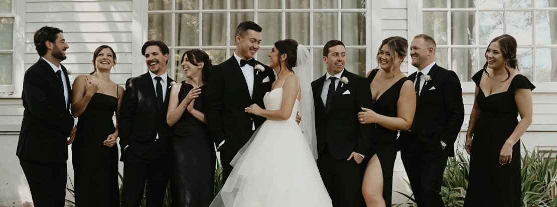 A joyful wedding party stands in front of a white building. The bride, in a voluminous white gown, and the groom, in a tuxedo, are surrounded by friends in black formal attire, all laughing and celebrating.