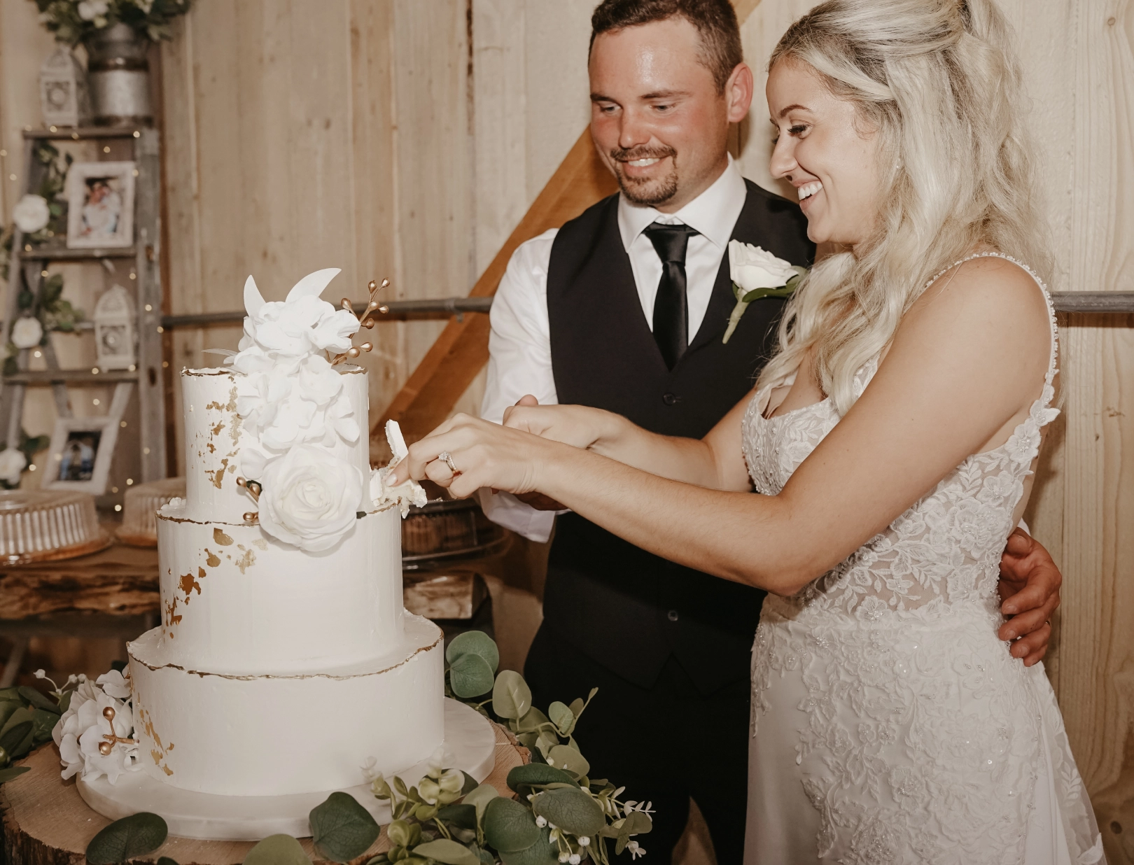 Bride and groom smiling while cutting a white, three-tiered wedding cake with floral decorations. The setting has a rustic, joyful vibe.