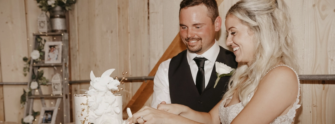 Bride and groom smiling while cutting a white, three-tiered wedding cake with floral decorations. The setting has a rustic, joyful vibe.
