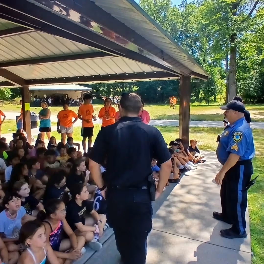 A group of children sitting under a pavilion listening to two police officers speaking during an outdoor event in a park.