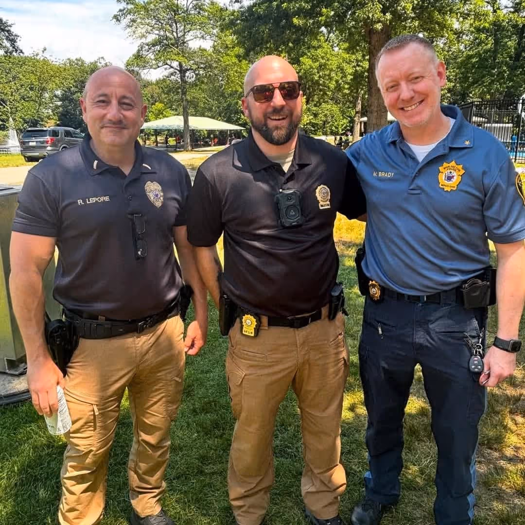 Three uniformed police officers standing outdoors on grass, smiling at the camera, with trees and park structures in the background.