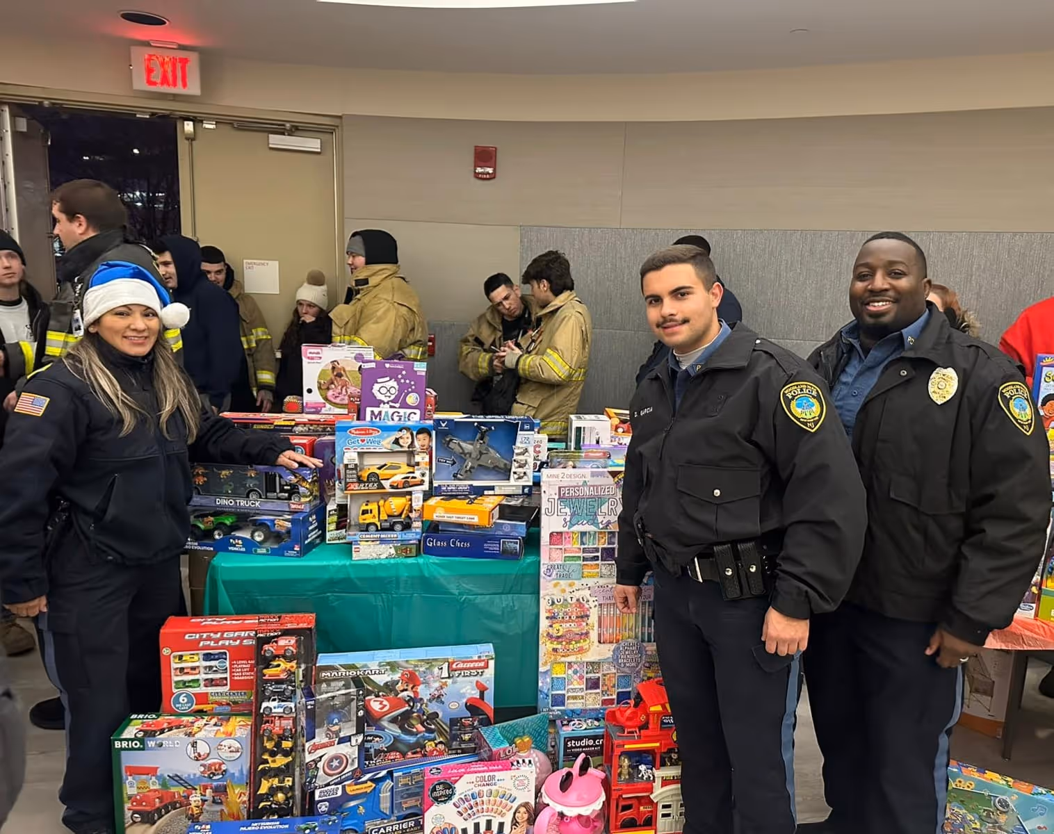 Three police officers standing and smiling by a table filled with various boxed toys in a room with people in the background.