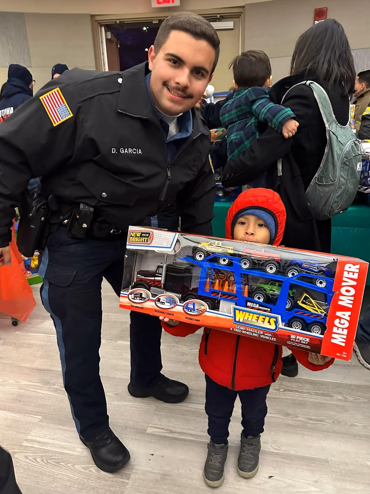 Police officer smiling and a child in a red jacket holding a toy car carrier playset indoors.