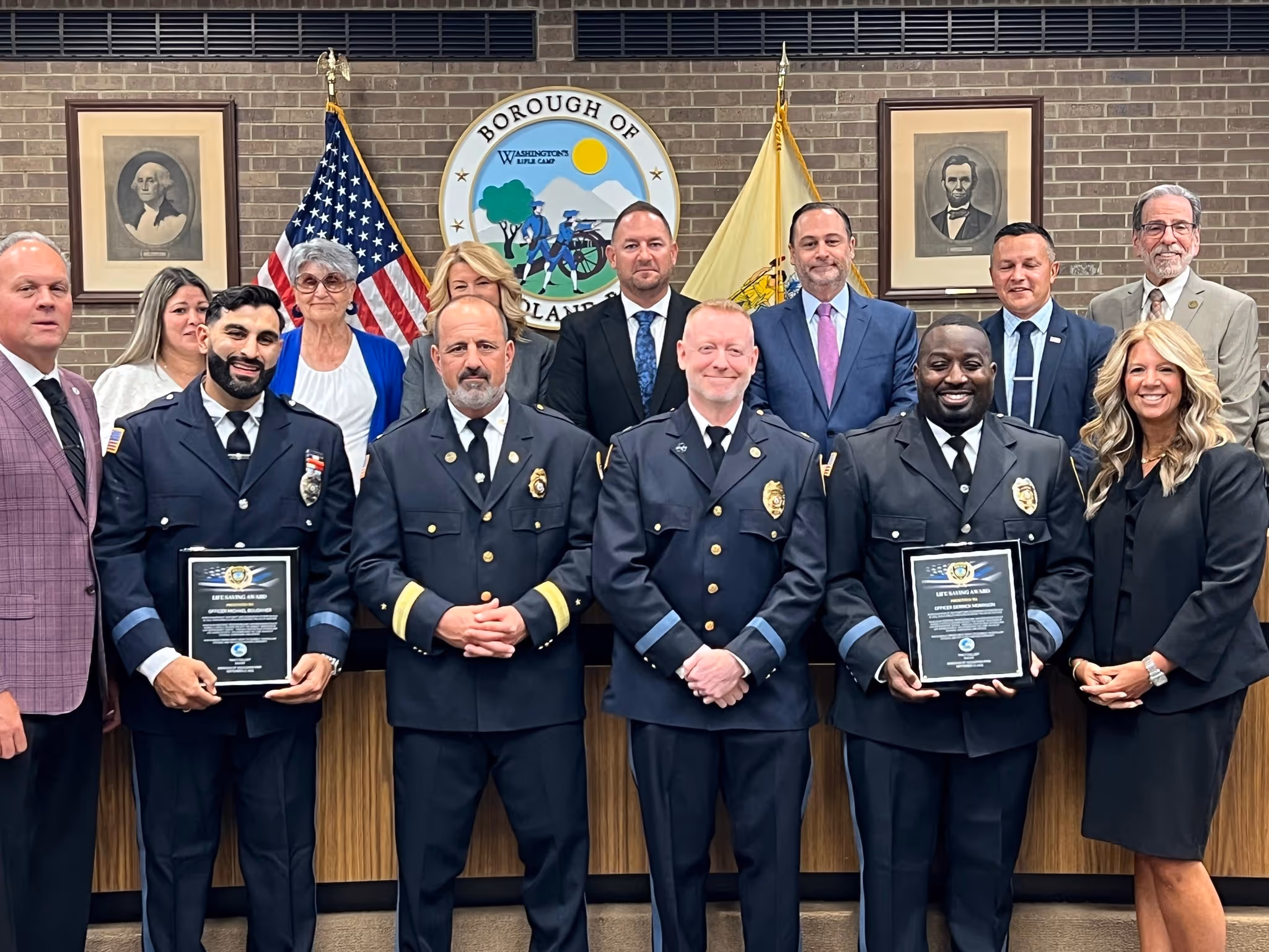 Group portrait of uniformed officers holding life-saving award plaques, standing with formally dressed officials in a government office.