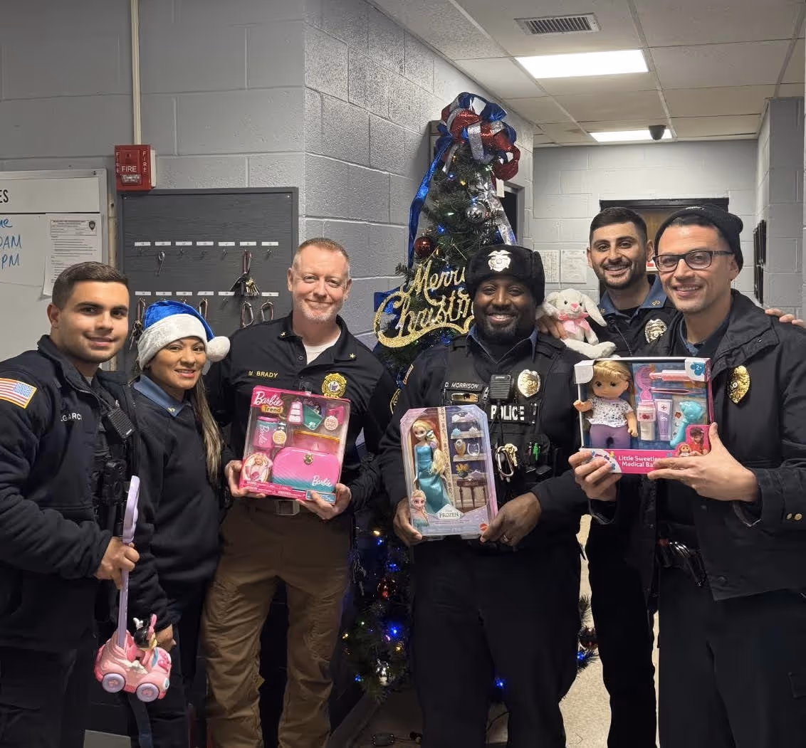 Six police officers smiling inside a station with a decorated Christmas tree in the background, each holding children's toys.