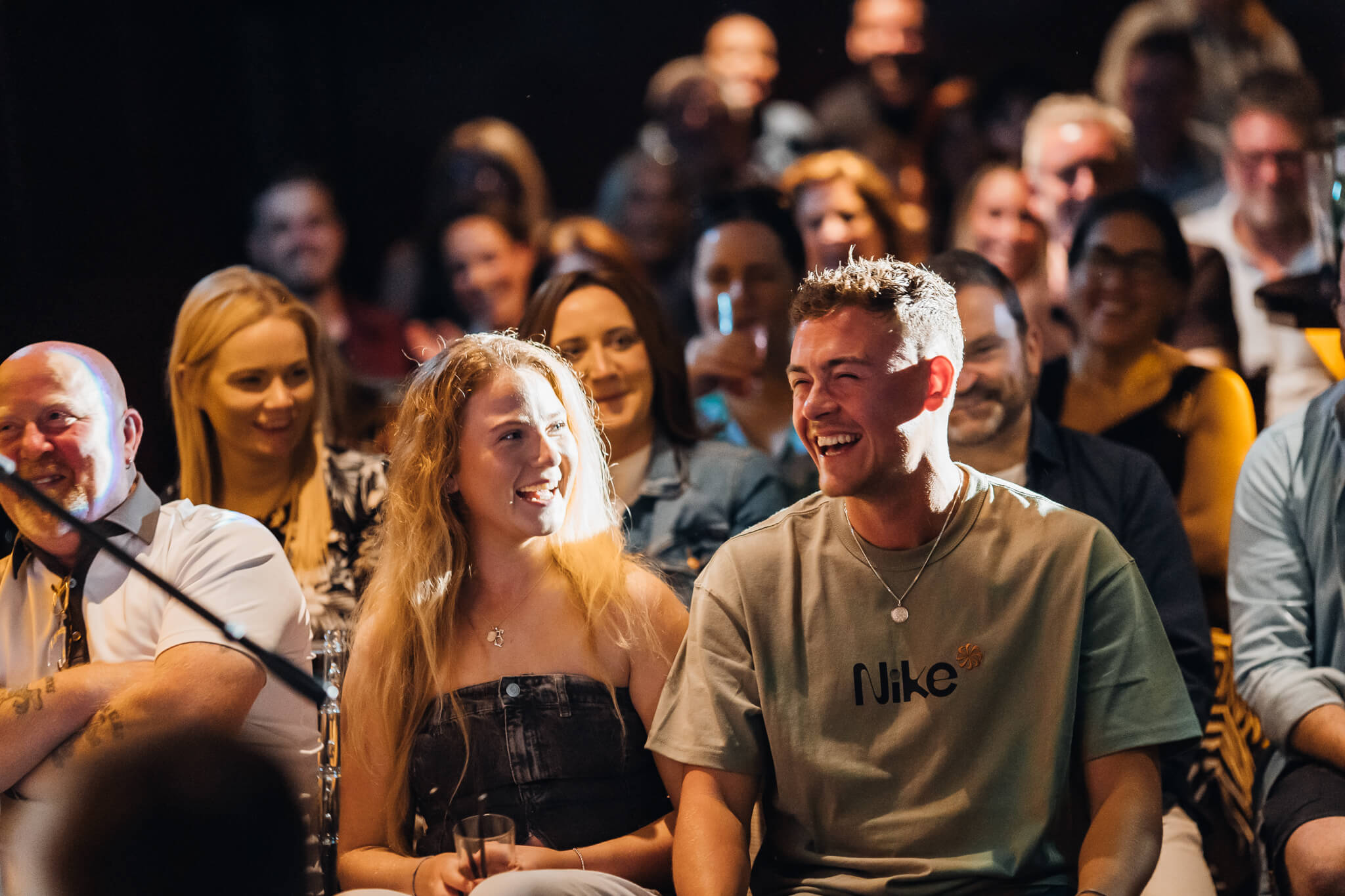 A lively audience at a comedy show, with several individuals laughing. Two people in the foreground, a young woman in a black top and a young man in a gray Nike shirt, are enjoying the performance. The setting appears to be Cavehill Comedy Club in Belfast.