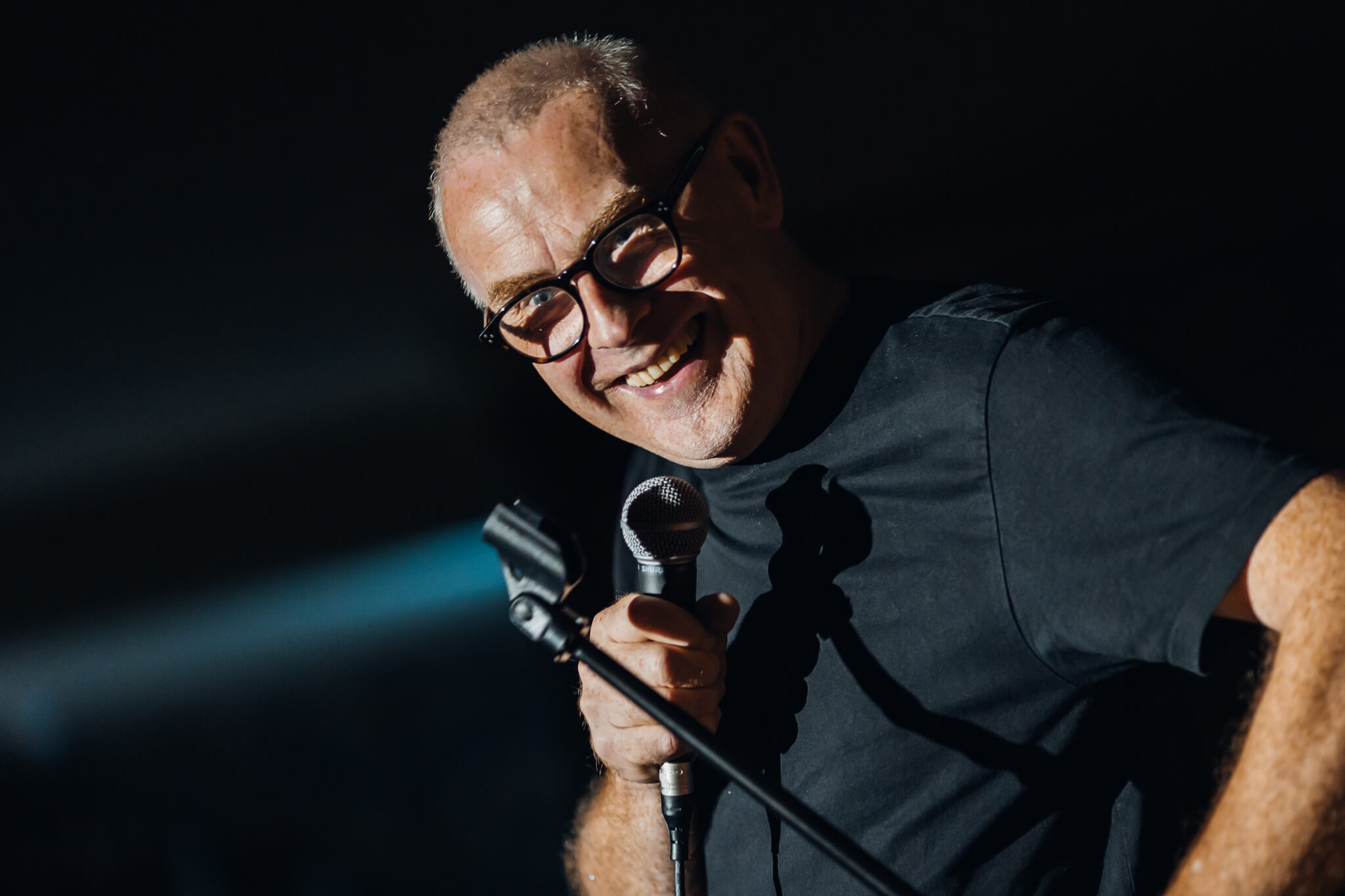 A smiling comedian with glasses holding a microphone on stage, illuminated by soft lighting. He appears engaged with the audience, possibly during a performance at Cavehill Comedy Club in Belfast.