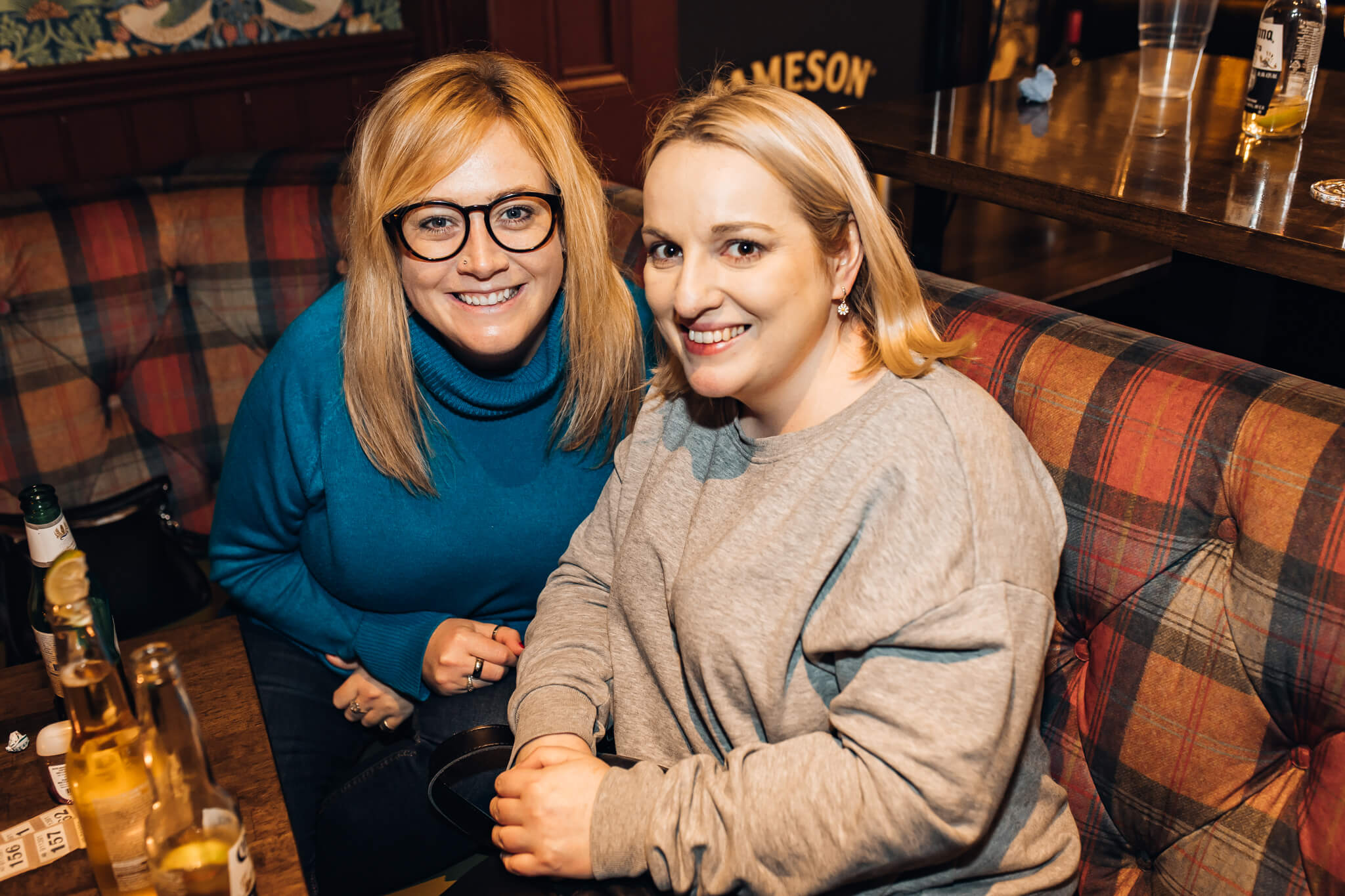 Two women sitting together at a table in a pub, smiling and posing for the camera. The setting features a plaid banquette and barware in the background. This image relates to social gatherings at Cavehill Comedy Club Belfast Thompsons.