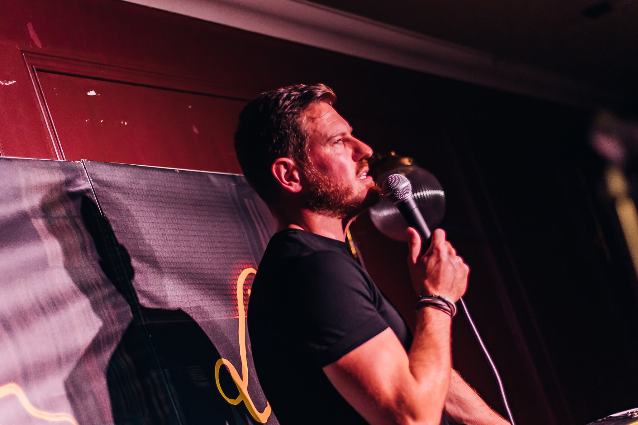 A comedian performing on stage at Cavehill Comedy Club in Belfast, holding a microphone and speaking passionately, with a black backdrop featuring colorful lights.