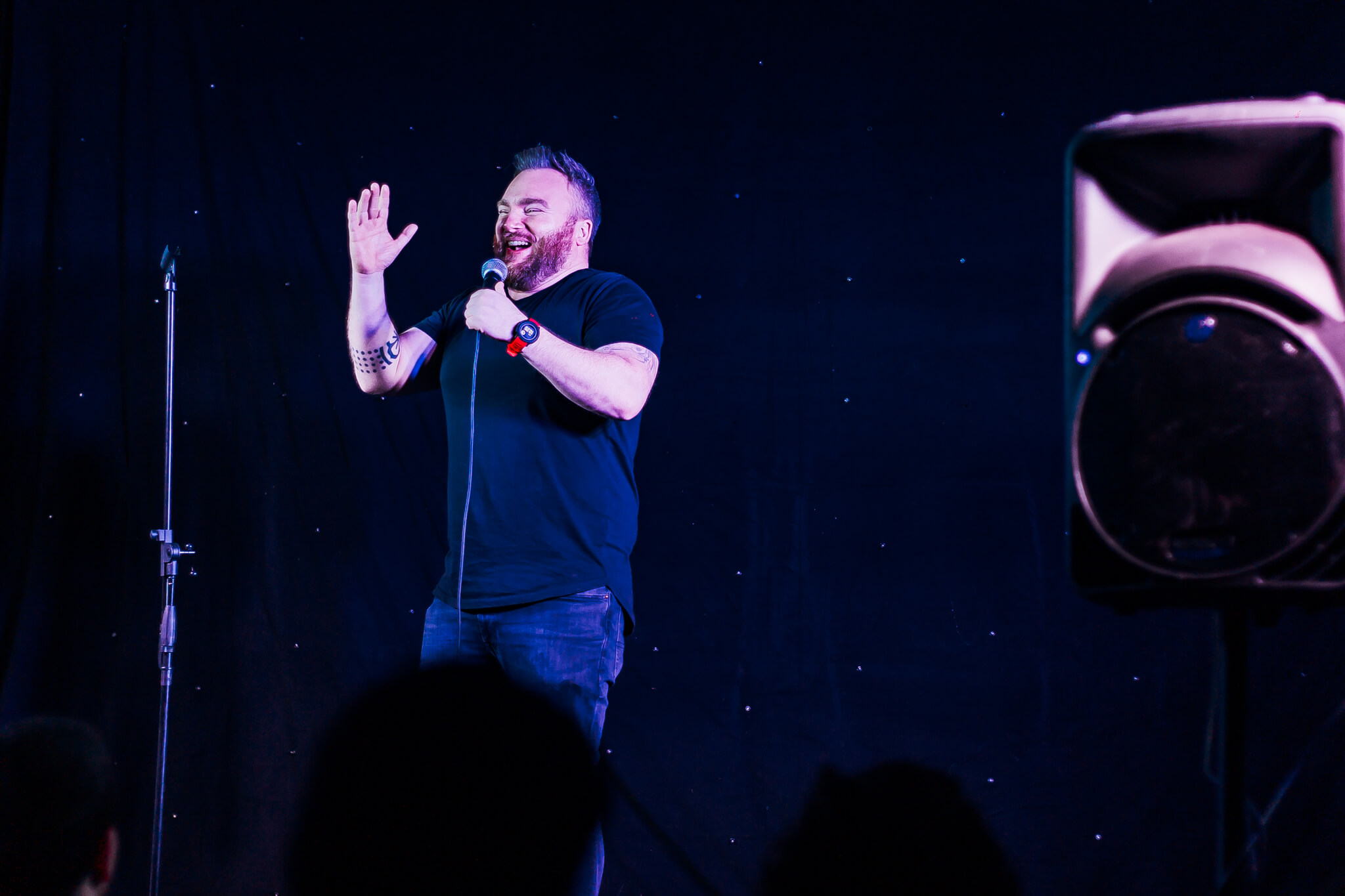 A comedian performing on stage at the Cavehill Comedy Club in Belfast. He is wearing a black shirt, holding a microphone, and gesturing with his hand while smiling. A speaker is visible to the right of the image.