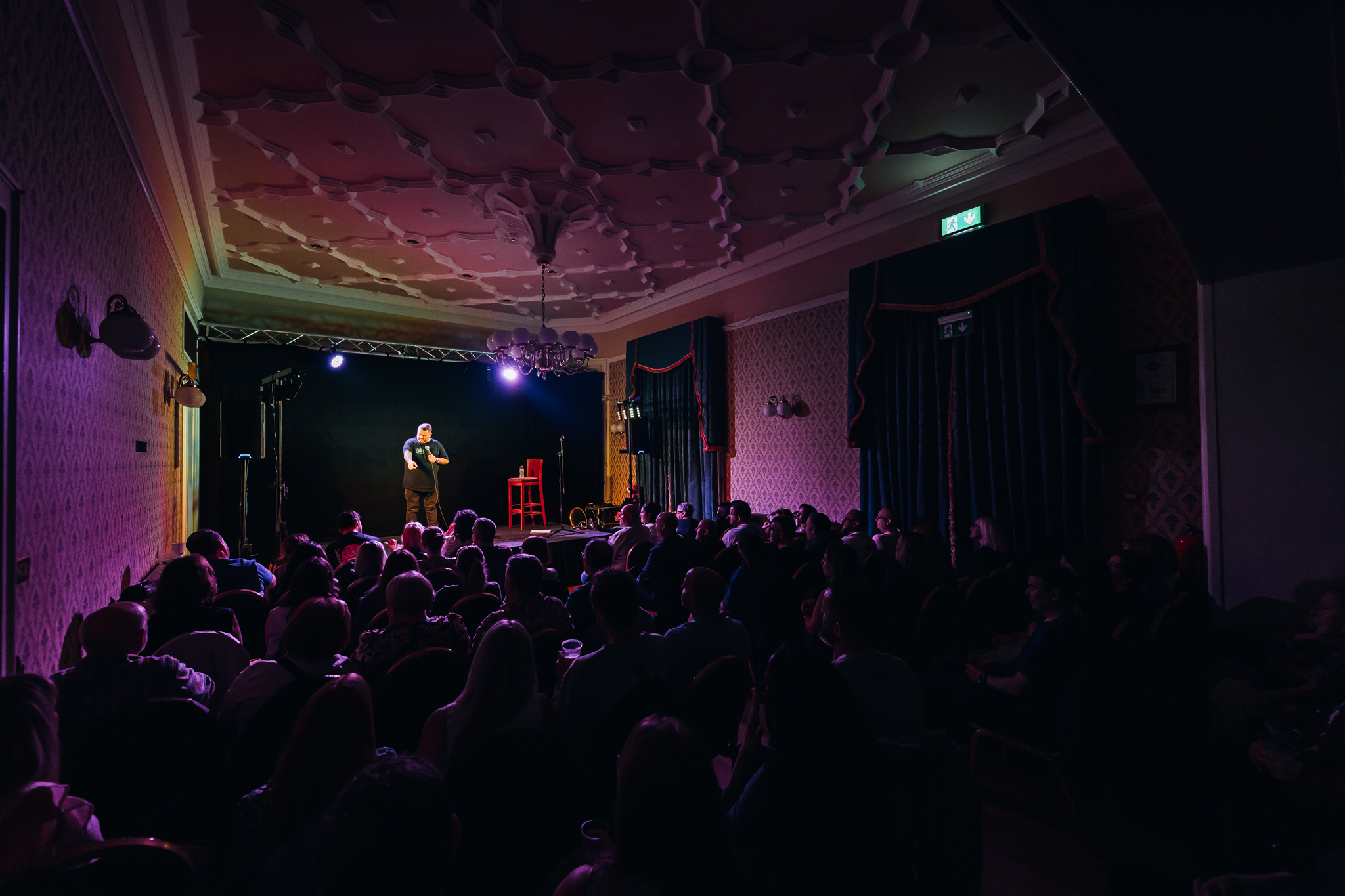 An audience at the Cavehill Comedy Club in Belfast enjoying a show, with a mix of expressions. In the foreground, a man in a plaid shirt is smiling, while others are engaged and attentive.