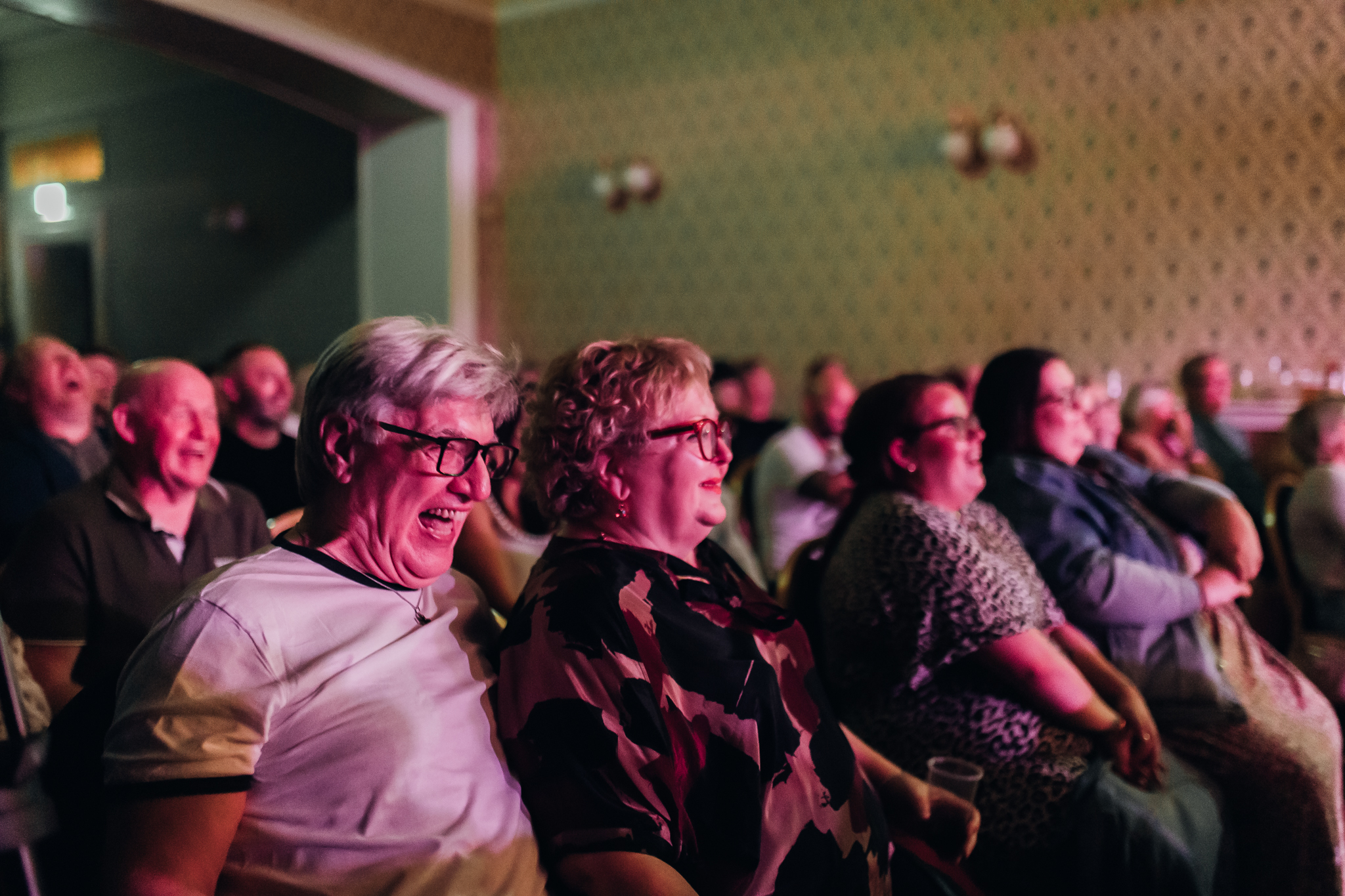 A comedian performing on stage at Cavehill Comedy Club in Belfast, holding a microphone and speaking passionately, with a black backdrop featuring colorful lights.